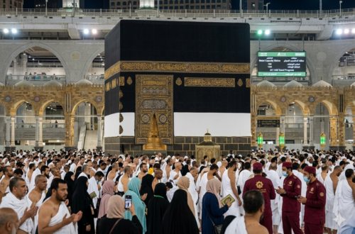 A panoramic view of the Holy Kaaba in Makkah during Hajj 2026, showing thousands of pilgrims in white Ihram performing Tawaf, with a digital information screen in the background displaying prayer times and pilgrim capacity updates.