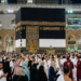 A panoramic view of the Holy Kaaba in Makkah during Hajj 2026, showing thousands of pilgrims in white Ihram performing Tawaf, with a digital information screen in the background displaying prayer times and pilgrim capacity updates.